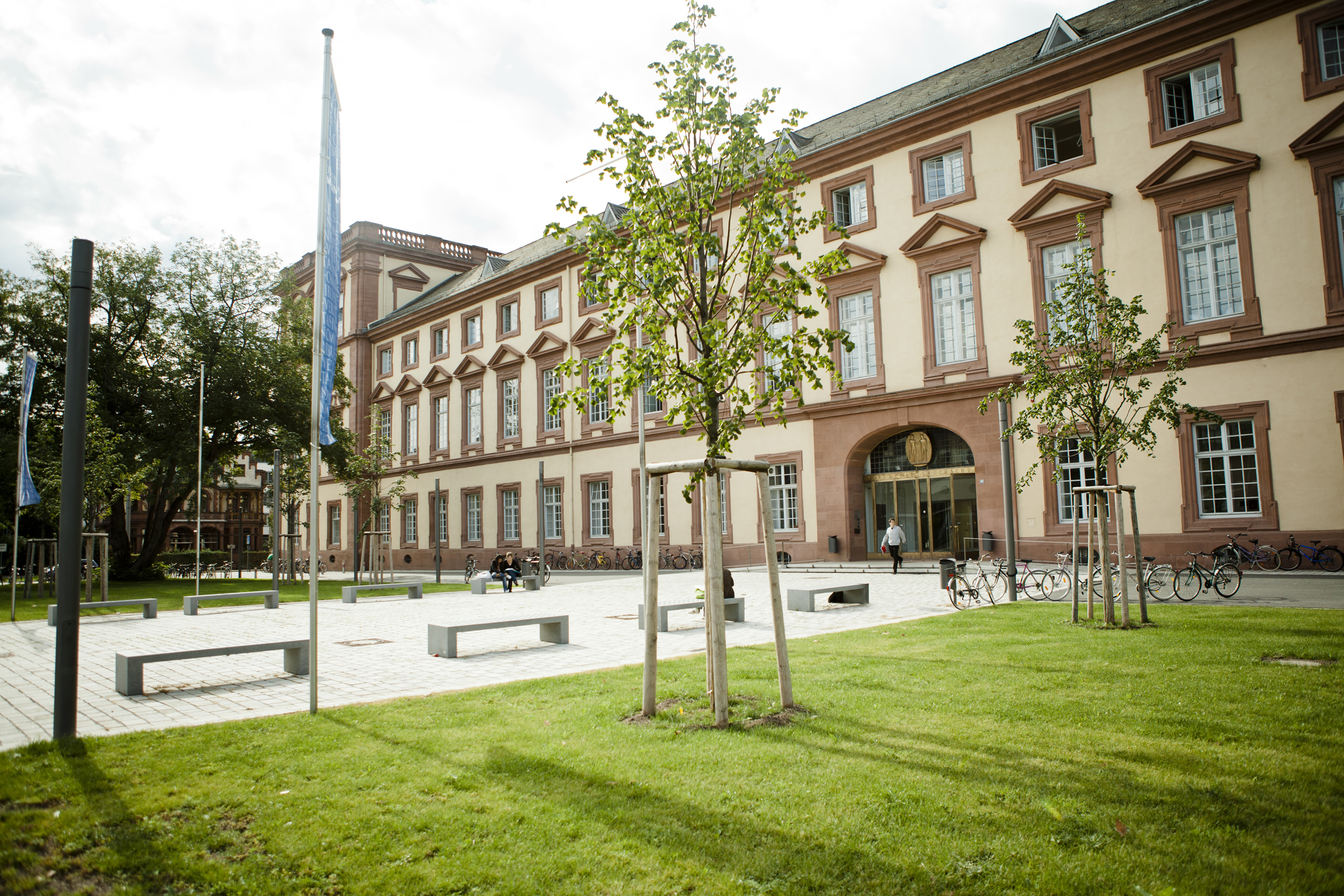 Exterior view of a University of Mannheim building with beige walls, red stone accents, and tall arched windows, facing a landscaped courtyard with young trees, benches, and a grassy lawn. A few people walk near the entrance, and bicycles are parked along the front of the building.